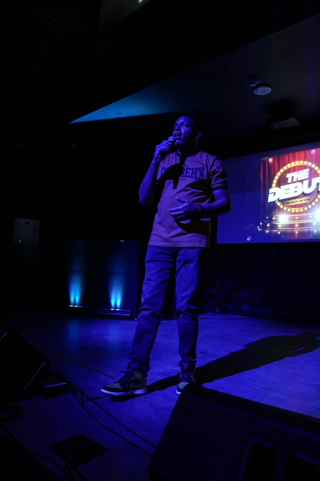 Chris Mayers hosting on stage with a microphone under blue lighting, The Debut screen behind him
