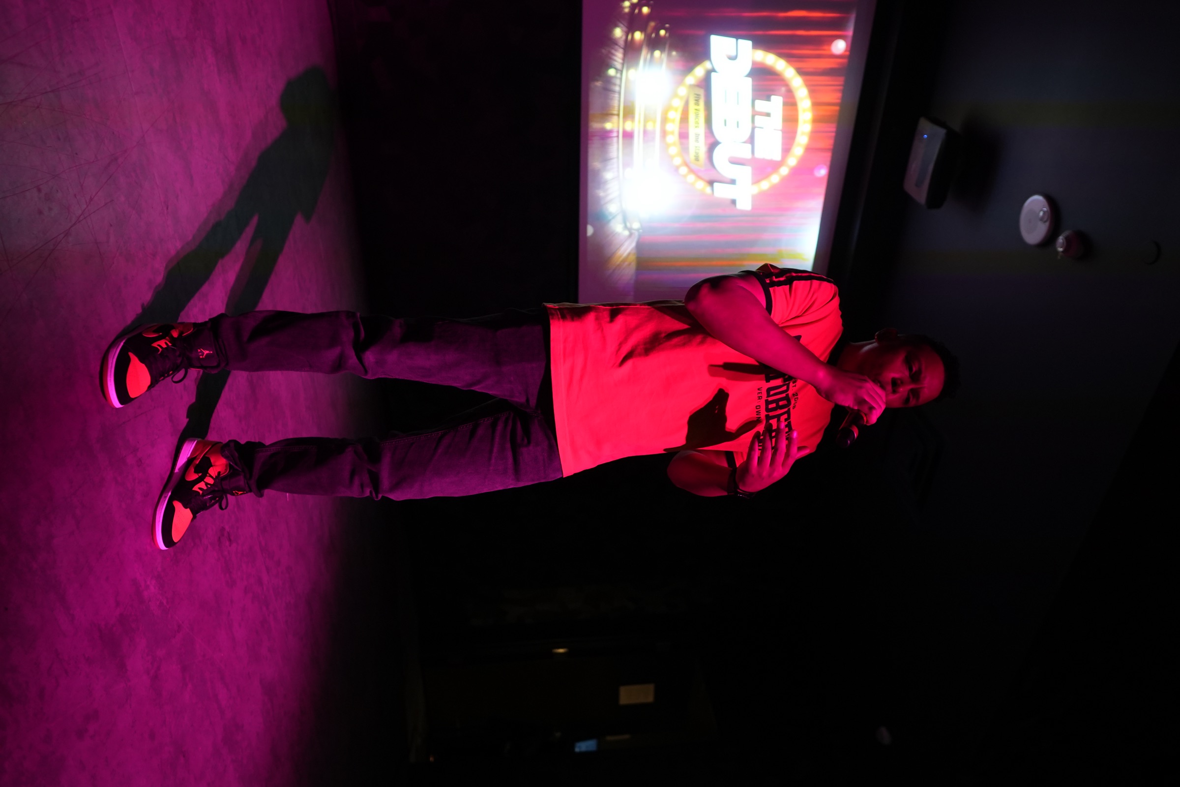 Chris Mayers hosting on the microphone under pink and red stage lighting at The Debut