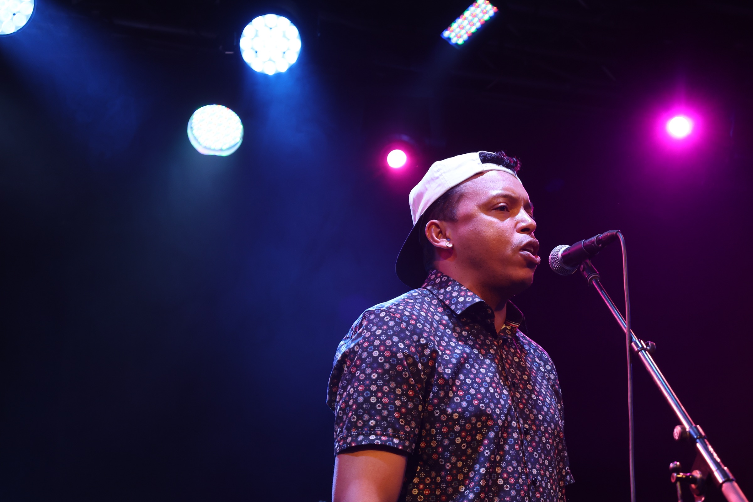Chris Mayers singing into a microphone on stage, stage lights behind him