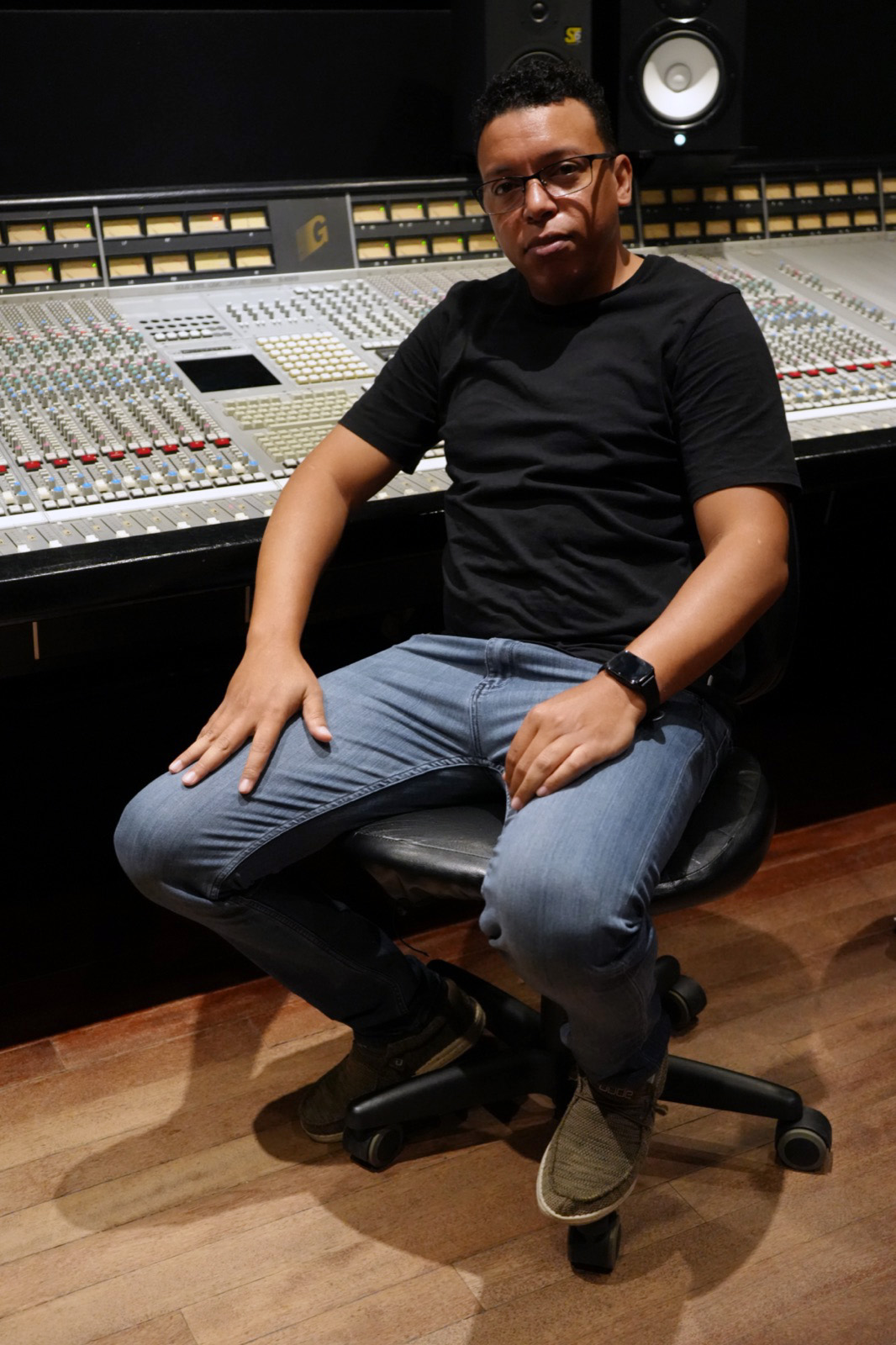 Chris Mayers seated at a large recording console with a studio monitor overhead